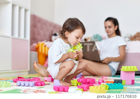 cute little baby girl playing with toys blocks and mother mother working on laptop from home cute little baby girl playing with toys blocks and mother mother working on laptop from home 117170810