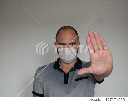 Stern looking man in medical mask shows open palm stop gesture on light background with copy space 117171056