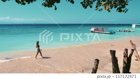 A woman is walking on the sandy beach next to a traditional wooden boat. The clear blue sky and gentle waves create a peaceful atmosphere. 117172871