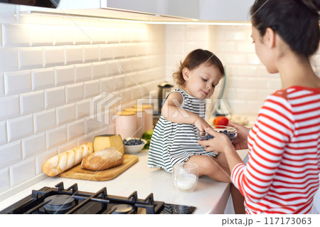 Little baby girl with her mother eating fresh blueberries in white kitchen Little baby girl with her mother eating fresh blueberries in white kitchen 117173063
