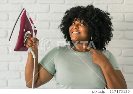 Young satisfied african american housekeeper woman wear mint t-shirt hold in hands on iron on brick wall background. Housework housekeeping concept 117173129