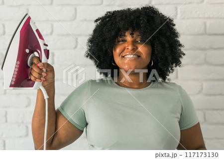 Young satisfied african american housekeeper woman wear mint t-shirt hold in hands on iron on brick wall background. Housework housekeeping concept 117173130