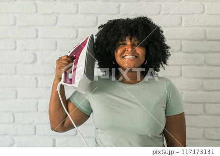 Young satisfied african american housekeeper woman wear mint t-shirt hold in hands on iron on brick wall background. Housework housekeeping concept 117173131