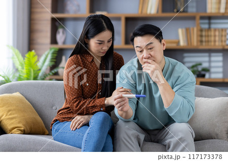 Asian couple sitting together on couch, examining pregnancy test. They look anxious and thoughtful, contemplating potential life changes. Shelves in background with plants, books, and decor. Asian couple sitting together on couch, examining pregnancy test. They look anxious and thoughtful, contemplating potential life changes. Shelves in background with plants, books, and decor. 117173378