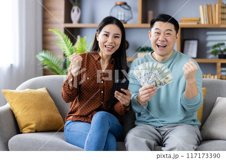 Asian couple celebrating financial success while sitting on couch holding smartphones and cash. They are happy and excited, showing positive expressions and gestures 117173390