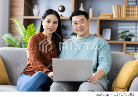 Asian couple sitting on couch using laptop at home. Man and woman smiling and looking at camera, enjoying time together. Concept of family, technology, internet 117173392