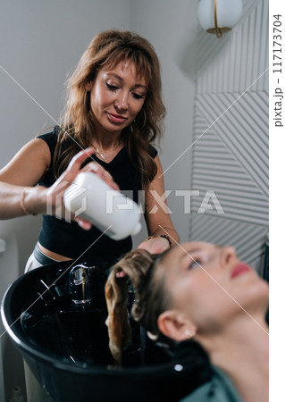 Vertical portrait of hairstylist foaming shampoo on long blonde hair of beautiful female client. Professional hairdresser washing head of young woman in beauty hairdressing salon. Vertical portrait of hairstylist foaming shampoo on long blonde hair of beautiful female client. Professional hairdresser washing head of young woman in beauty hairdressing salon. 117173704