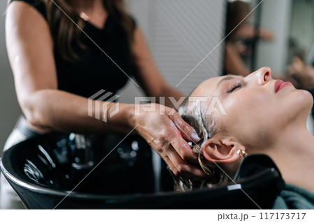 Close-up cropped shot of unrecognizable hairstylist foaming shampoo on long blonde hair of beautiful female client. Closeup of hairdresser washing head of young woman in beauty hairdressing salon. Close-up cropped shot of unrecognizable hairstylist foaming shampoo on long blonde hair of beautiful female client. Closeup of hairdresser washing head of young woman in beauty hairdressing salon. 117173717
