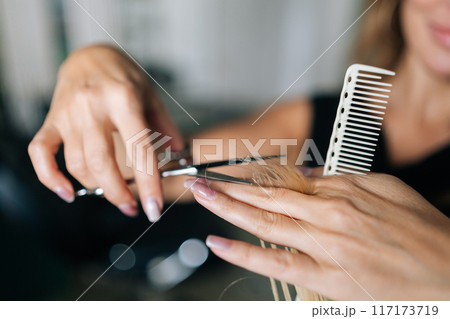 Close-up hands of unrecognizable female hairdresser making stylish haircut with professional scissors to woman client with long blonde hair. Hairstylist cutting lady hair with barber scissors. 117173719