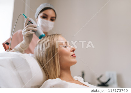 Closeup side view of attractive blonde female patient having hair light therapy with modern innovation apparatus. Trichologist performing hair treatment procedure with spectrum phototherapy device. 117173740