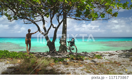 Tourist woman in swimsuit stands by tree on a tropical beach in French Polynesia. Turquoise ocean water, clear blue sky in warm, sunny day, tranquil and peaceful atmosphere. Summer holiday vacation Tourist woman in swimsuit stands by tree on a tropical beach in French Polynesia. Turquoise ocean water, clear blue sky in warm, sunny day, tranquil and peaceful atmosphere. Summer holiday vacation 117173867