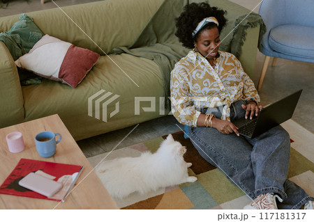 Smiling woman sitting on couch and typing on laptop with fluffy cat lying on floor beside coffee table with books and cups adding cozy atmosphere 117181317