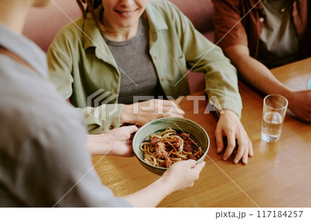 Person providing a bowl of Asian noodles to two people seated at table. Simple and cozy setup with clear focus on the food 117184257