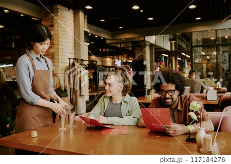 Group of diners sitting at wooden table in cozy restaurant ambiance while waitress pouring water. Warm lighting and casual atmosphere create inviting dining experience Group of diners sitting at wooden table in cozy restaurant ambiance while waitress pouring water. Warm lighting and casual atmosphere create inviting dining experience 117184276