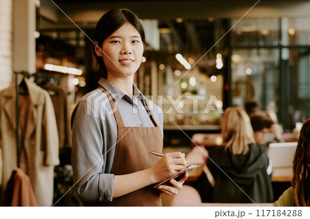 Smiling Asian waitress in apron holding notepad in busy cafe, engaging with customers. Background shows bustling cafe interior with patrons seated at tables and bar area Smiling Asian waitress in apron holding notepad in busy cafe, engaging with customers. Background shows bustling cafe interior with patrons seated at tables and bar area 117184288