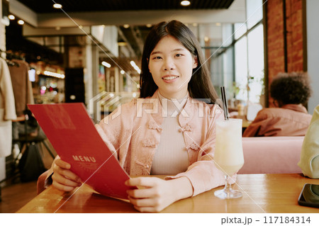 Portrait of Asian woman reading menu while seated in cozy cafe, enjoying refreshing drink with visible patrons and warm decor in background 117184314