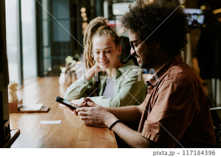 Smiling people relaxing in cozy cafe and using smartphones while enjoying coffee. Modern setting with natural light and friendly atmosphere enhancing experience 117184396