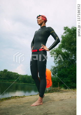 Energetic swimmer in black wetsuit and red cap, with orange buoy, dashes into lake, preparing to marathon. Energetic swimmer in black wetsuit and red cap, with orange buoy, dashes into lake, preparing to marathon. 117186352