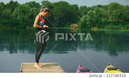 Female swimmer with warms up on dock by calm lake, surrounded by nature. Her wetsuit and gear indicate readiness for water sports. 117186410