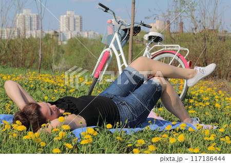 The girl is resting on the spring meadow. Lies on the bedspread. Nearby is a bike. Dandelions are blooming, young grass is growing. 117186444