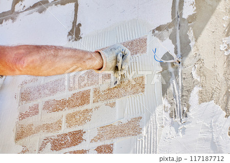 Brick cladding, also called brick veneer is attached to exterior of house made of aerated concrete blocks, construction worker uses tile adhesive. Brick cladding, also called brick veneer is attached to exterior of house made of aerated concrete blocks, construction worker uses tile adhesive. 117187712