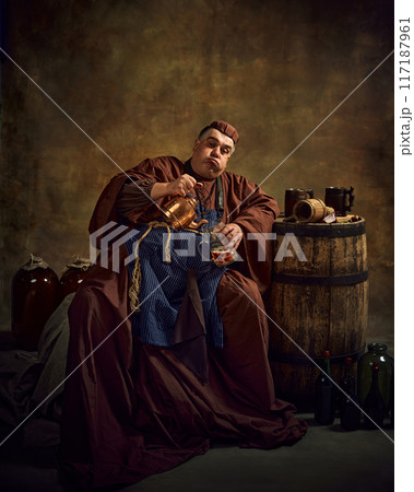 Medieval monk in period clothes pouring beer into mug from kettle, sitting in dimly lit cellar with wooden barrel and beer mug on top. Oktoberfest Medieval monk in period clothes pouring beer into mug from kettle, sitting in dimly lit cellar with wooden barrel and beer mug on top. Oktoberfest 117187961