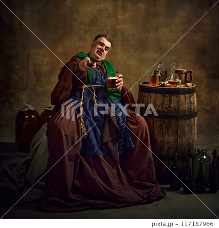Sport fan supporting soccer team. Emotional monk in period attire watching game, drinking beer gain dimly lit vintage background. Sport fan supporting soccer team. Emotional monk in period attire watching game, drinking beer gain dimly lit vintage background. 117187966