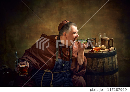 Man, monk in period attire enjoying sandwich with fish and drinking ale, against vintage background with wooden barrel with various beer mugs on top Man, monk in period attire enjoying sandwich with fish and drinking ale, against vintage background with wooden barrel with various beer mugs on top 117187969
