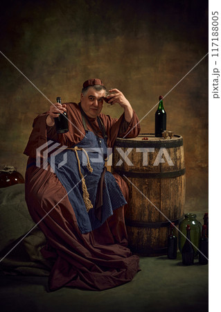 Hangover. Medieval man, monk in traditional attire leaning glass with beverage to head, leaning on wooden barrel with bottle. Cheers Hangover. Medieval man, monk in traditional attire leaning glass with beverage to head, leaning on wooden barrel with bottle. Cheers 117188005