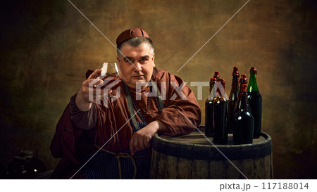 Man, medieval monk in robe raising glass filled with brown liquid, beer, ale, cognac against vintage background. Traditions of abbey brewing. Man, medieval monk in robe raising glass filled with brown liquid, beer, ale, cognac against vintage background. Traditions of abbey brewing. 117188014