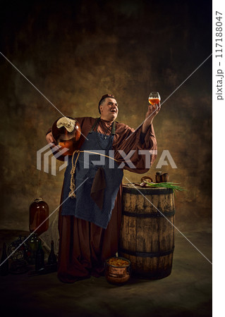 Medieval monk, man in period clothes with beer barrel and pint raising glass of ale against vintage background. Ancient craft of monastic ale production. Medieval monk, man in period clothes with beer barrel and pint raising glass of ale against vintage background. Ancient craft of monastic ale production. 117188047