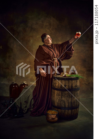 Dedication to abbey brewing through the ages. Medieval monk raising glass with freshly brewed ale, beer against stage background. Dedication to abbey brewing through the ages. Medieval monk raising glass with freshly brewed ale, beer against stage background. 117188054