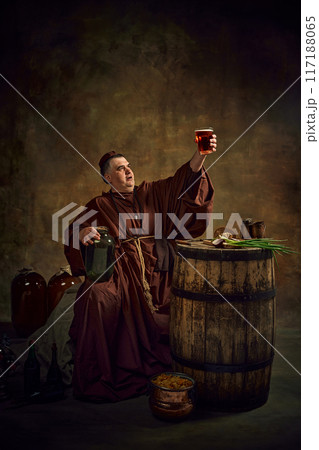 Man, medieval monk lifting glass of beer, pint of ale against dark vintage background. Rich legacy of abbey brewing traditions 117188065
