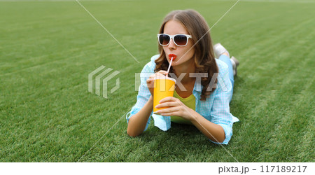 Happy young woman with cup of coffee, girl drinking fresh juice lying on green grass in summer park 117189217