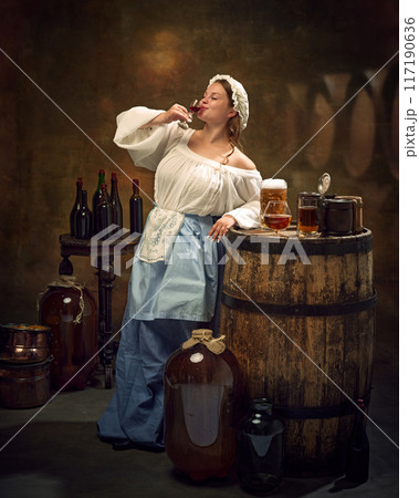 Woman in traditional attire drinking alcohol drink from glass, surrounded by barrels, bottles, and various brews at vintage pub. 117190636