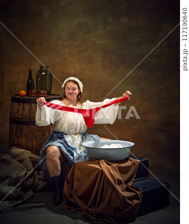 Young chubby medieval woman washing clothes. Lady in old-fashion clothes looks proudly on red oversized pants against vintage background. 117190640