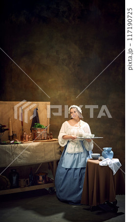 Young chubby woman, maiden, in historical dress holds tray, as waitress surrounded by vintage kitchenware and herbs. 117190725