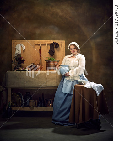 Portrait of young woman in traditional attire, holding blue pitcher, stands by rustic kitchen setup against vintage background. 117190733