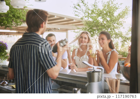 Group of friends relaxing with cocktails at outdoor bar, engaging in lively conversation with male bartender. Outdoor drinks Group of friends relaxing with cocktails at outdoor bar, engaging in lively conversation with male bartender. Outdoor drinks 117190763