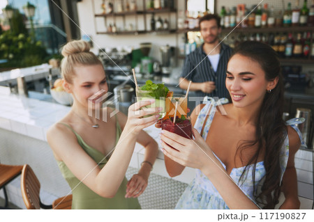 Cheers. Two smiling beautiful women clinking glasses with cocktails in modern bar settings, with bartender on background 117190827