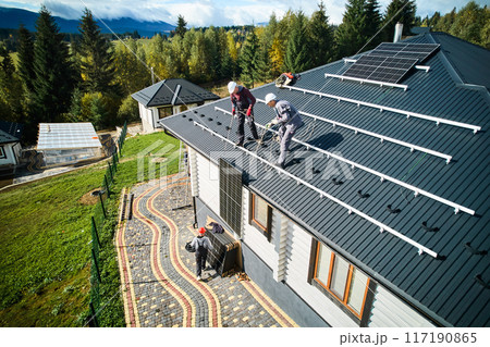 Men installers installing solar panel system on roof of house. Roofers in helmets lifting up photovoltaic solar module with help of ropes outdoors. Concept of alternative and renewable energy. 117190865