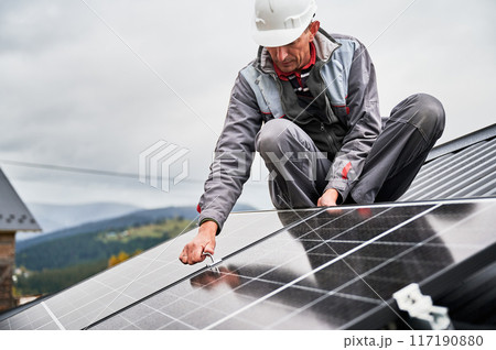 Man worker mounting photovoltaic solar panels on roof of house. Engineer in helmet installing solar module system with help of hex key. Concept of alternative, renewable energy. Man worker mounting photovoltaic solar panels on roof of house. Engineer in helmet installing solar module system with help of hex key. Concept of alternative, renewable energy. 117190880