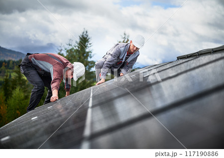 Mounters installing photovoltaic solar panels on roof of house. Men engineers in helmets building solar module system with help of hex key. Concept of alternative, renewable energy. 117190886