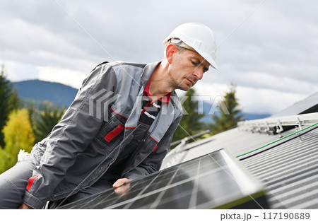 Man builder mounting photovoltaic solar moduls on roof of house. Close up engineer in helmet looking on evenness of solar panel during installing. Concept of alternative and renewable energy. Man builder mounting photovoltaic solar moduls on roof of house. Close up engineer in helmet looking on evenness of solar panel during installing. Concept of alternative and renewable energy. 117190889