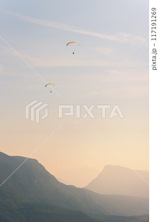 Two paragliders in the air fly over the mountains at summer sunrise. Two paragliders in the air fly over the mountains at summer sunrise. 117191269