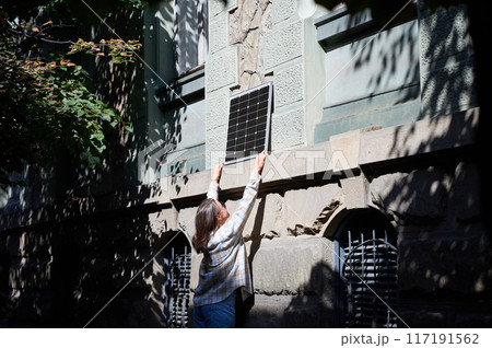 Woman holding photovoltaic solar panel in front of historical building, wearing plaid shirt and sunglasses. Concept of integration of sustainable renewable energy sources into old architecture. 117191562