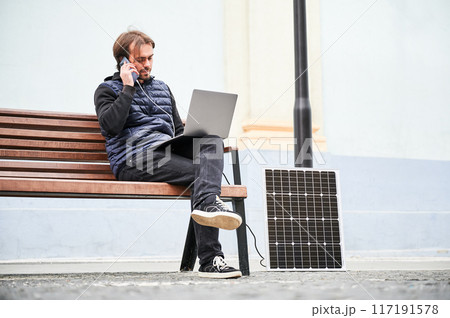 Man sits on wooden bench, working on laptop and smartphone, both connected to solar panel. Urban setting and casual attire highlight modern, sustainable, mobile workspace. Man sits on wooden bench, working on laptop and smartphone, both connected to solar panel. Urban setting and casual attire highlight modern, sustainable, mobile workspace. 117191578
