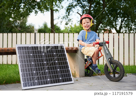 Young happy boy sits on balance bike next to solar panel and wooden bench, wearing red helmet, smiling brightly at camera. Background features white fence, trees. Concept of green energy generation. 117191579