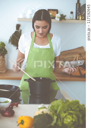 An attractive young dark-haired woman preparing soup by new keto recipe while standing and smiling in the kitchen. Cooking and householding concepts 117192645