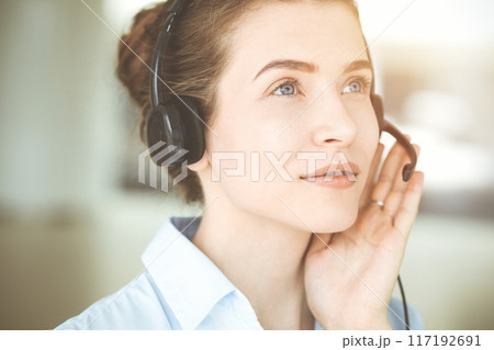 Beautiful female specialist with headphones helping customers online and smiling charmingly in sunny call center office. Business people concept 117192691